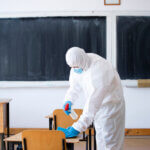 The image depicts a person wearing a PPE and sanitizing the school using EN 14347 tested disinfectants.