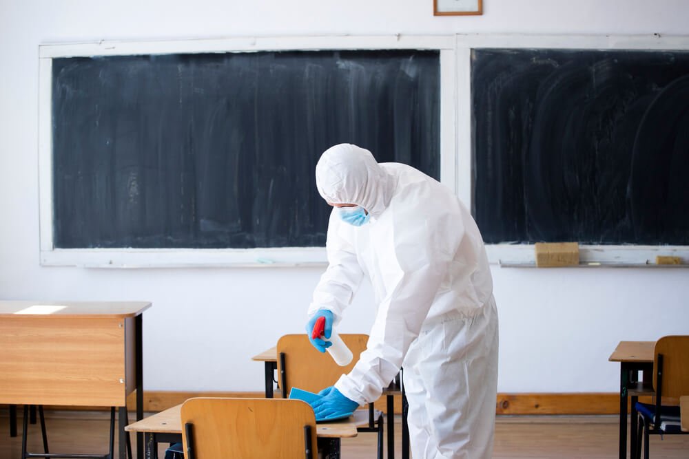 The image depicts a person wearing a PPE and sanitizing the school using EN 14347 tested disinfectants.