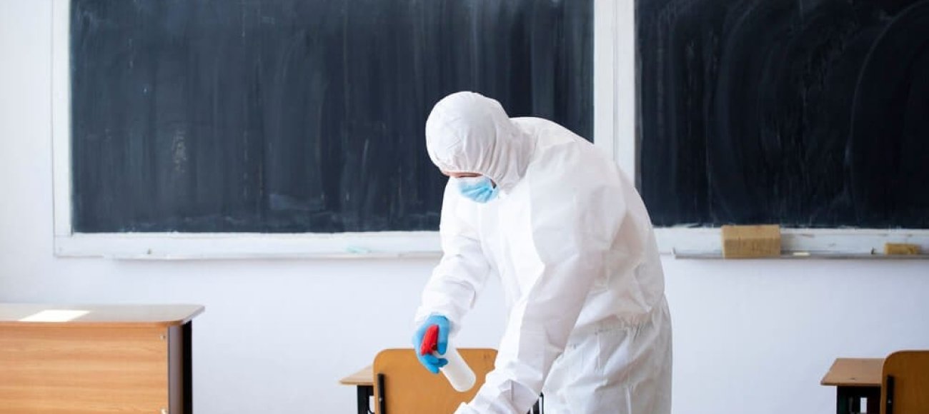 The image depicts a person wearing a PPE and sanitizing the school using EN 14347 tested disinfectants.